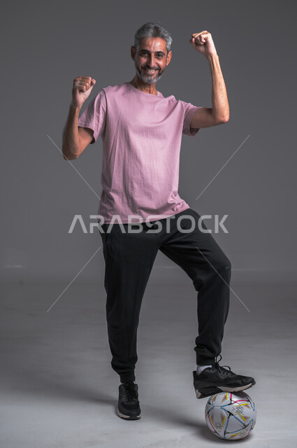 لاعب كرة قدم محترف ،ممارسة تمرينات رياضة ،  ايماءات الوجه واليدين تدل على الفوز ، بورتريه لرجل عربي خليجي سعودي يرتدي الزي الرياضي ويضع الكره تحت قدمه ، صوره لكامل الجسم ، خلفيه رماديه