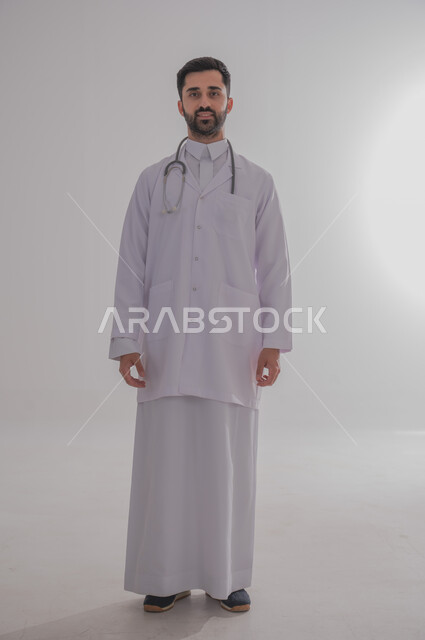 العمل في القطاع الصحي في السعودية، النظر للكاميرا بإيماءات الثقة بالنفس والسرور, الفحص الطبي وتقديم الخدمات الصحية للمرضى, بورتريه لطبيب عربي خليجي سعودي مبتسم يرتدي الثوب التقليدي والمعطف الطبي يضع السماعة الطبية حول عنقه، الجسم بالطول الكامل، خلفية رمادية