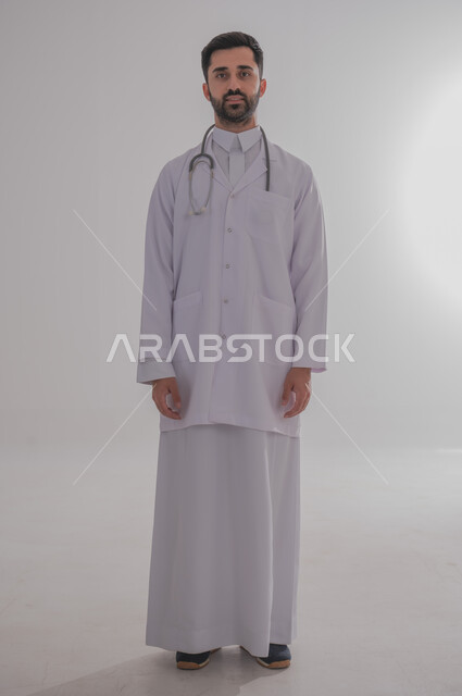 العمل في القطاع الصحي في السعودية، النظر للكاميرا بإيماءات الثقة بالنفس والسرور, الفحص الطبي وتقديم الخدمات الصحية للمرضى, بورتريه لطبيب عربي خليجي سعودي مبتسم يرتدي الثوب التقليدي والمعطف الطبي يضع السماعة الطبية حول عنقه، الجسم بالطول الكامل، خلفية رمادية