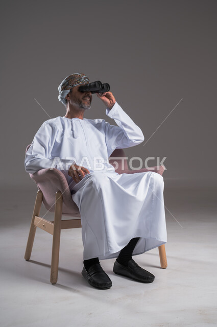 استعمال المنظار للكشف عن الاشياء البعيدة وتقريبها,  بورتريه لرجل عربي خليجي عماني يرتدي الكمه والدشداشة يجلس على كرسي مريح يحمل بيده المنظار ثنائي العينين وينظر الى شيء ما، حب الاستكشاف والمغامرة، إيماءات الاستمتاع والسرور، خلفية رمادية