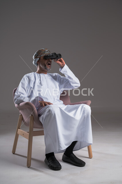 استعمال المنظار للكشف عن الاشياء البعيدة وتقريبها,  بورتريه لرجل عربي خليجي عماني يرتدي الكمه والدشداشة يجلس على كرسي مريح يحمل بيده المنظار ثنائي العينين وينظر الى شيء ما، حب الاستكشاف والمغامرة، إيماءات الاستمتاع والسرور، خلفية رمادية