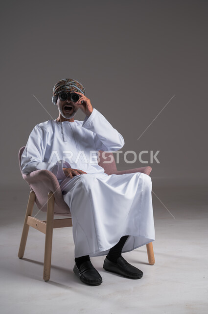 استعمال المنظار للكشف عن الاشياء البعيدة وتقريبها,  بورتريه لرجل عربي خليجي عماني يرتدي الكمه والدشداشة يجلس على كرسي مريح يحمل بيده المنظار ثنائي العينين وينظر الى شيء ما، حب الاستكشاف والمغامرة، إيماءات الاستمتاع والسرور، خلفية رمادية