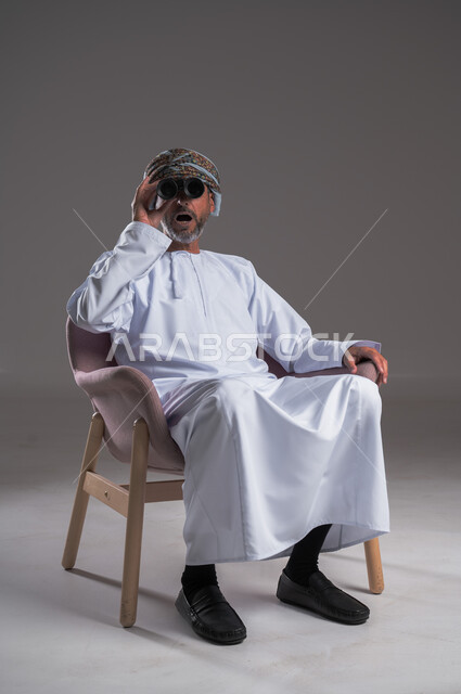 استعمال المنظار للكشف عن الاشياء البعيدة وتقريبها,  بورتريه لرجل عربي خليجي عماني يرتدي الكمه والدشداشة يجلس على كرسي مريح يحمل بيده المنظار ثنائي العينين وينظر الى شيء ما، حب الاستكشاف والمغامرة، إيماءات الاستمتاع والسرور، خلفية رمادية