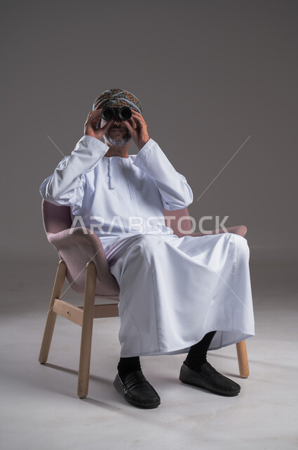 استعمال المنظار للكشف عن الاشياء البعيدة وتقريبها,  بورتريه لرجل عربي خليجي عماني يرتدي الكمه والدشداشة يجلس على كرسي مريح يحمل بيده المنظار ثنائي العينين وينظر الى شيء ما، حب الاستكشاف والمغامرة، إيماءات الاستمتاع والسرور، خلفية رمادية