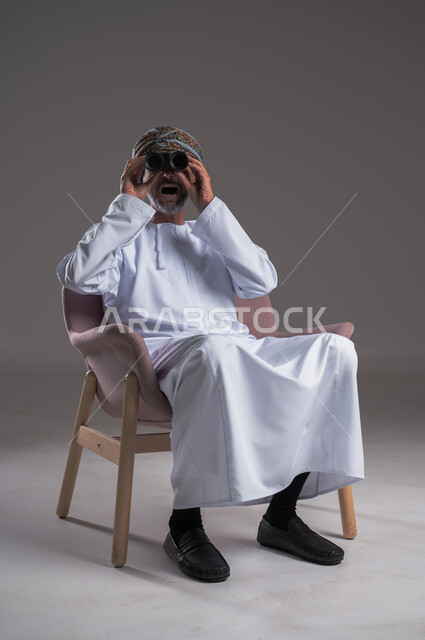 استعمال المنظار للكشف عن الاشياء البعيدة وتقريبها,  بورتريه لرجل عربي خليجي عماني يرتدي الكمه والدشداشة يجلس على كرسي مريح يحمل بيده المنظار ثنائي العينين وينظر الى شيء ما، حب الاستكشاف والمغامرة، إيماءات الاستمتاع والسرور، خلفية رمادية