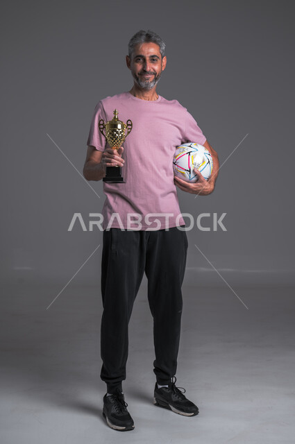 Hobby of playing ball, gestures indicating happiness, professional football player, portrait of a Saudi Arabian Gulf man wearing sportswear holding ball and winning cup, full body photo, gray background