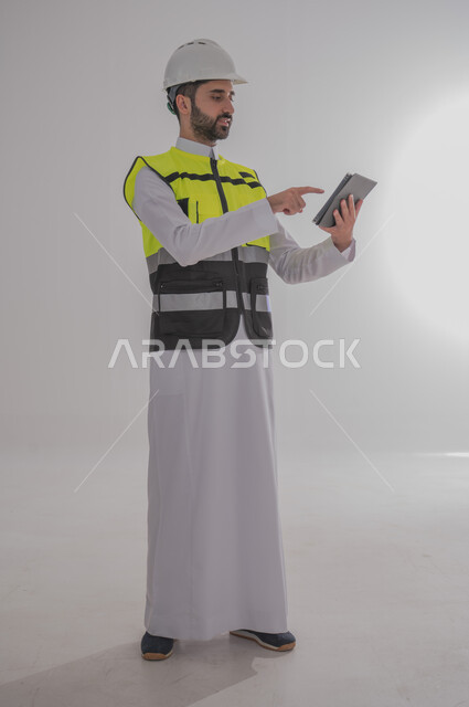 مهن ووظائف سعودية للشباب، الإعمار بسواعد أبناء الوطن، مفهوم الهندسة والانشاءات المعمارية، بورتريه لمهندس عربي خليجي سعودي يرتدي الثوب التقليدي والسترة الخاصة وخوذة الحماية يقف ويمسك بدفتر الملاحظات ويتابع العمل حسب المخطط، الجسم بالطول الكامل، خلفية رمادية