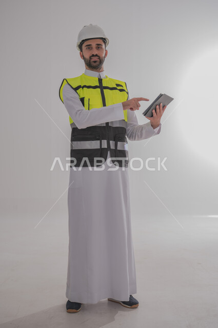 مهن ووظائف سعودية للشباب، الإعمار بسواعد أبناء الوطن، مفهوم الهندسة والانشاءات المعمارية، بورتريه لمهندس عربي خليجي سعودي يرتدي الثوب التقليدي والسترة الخاصة وخوذة الحماية يقف ويمسك بدفتر الملاحظات ويتابع العمل حسب المخطط، الجسم بالطول الكامل، خلفية رمادية