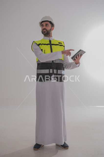 مهن ووظائف سعودية للشباب، الإعمار بسواعد أبناء الوطن، مفهوم الهندسة والانشاءات المعمارية، بورتريه لمهندس عربي خليجي سعودي يرتدي الثوب التقليدي والسترة الخاصة وخوذة الحماية يقف ويمسك بدفتر الملاحظات ويتابع العمل حسب المخطط، الجسم بالطول الكامل، خلفية رمادية