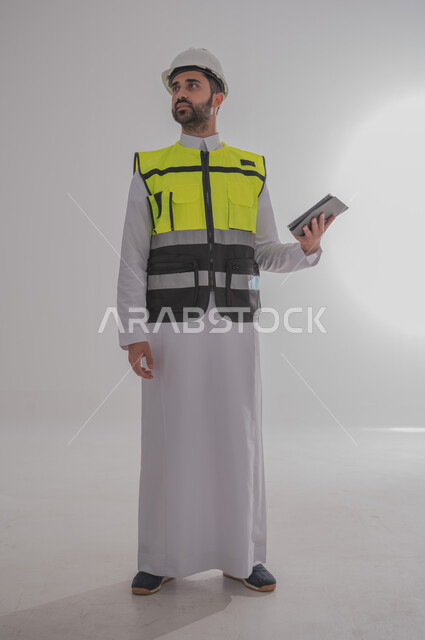 مهن ووظائف سعودية للشباب، الإعمار بسواعد أبناء الوطن، مفهوم الهندسة والانشاءات المعمارية، بورتريه لمهندس عربي خليجي سعودي يرتدي الثوب التقليدي والسترة الخاصة وخوذة الحماية يقف ويمسك بدفتر الملاحظات ويتابع العمل حسب المخطط، الجسم بالطول الكامل، خلفية رمادية