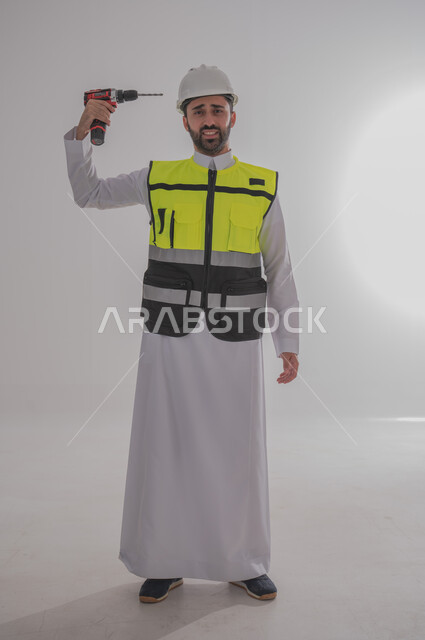 العمل في القطاع الهندسي، الإعمار بسواعد أبناء الوطن، مهن ووظائف سعودية، مفهوم الهندسة والانشاءات المعمارية، بورتريه لمهندس عربي خليجي سعودي يرتدي الثوب التقليدي والسترة الخاصة وخوذة الحماية يقف بشكل مستقيم، استخدام الأدوات والمعدات الخطيرة، خلفية رمادية