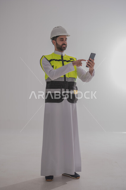 استخدام التقنيات والتكنولوجيا الحديثة في القطاع الهندسي، الإعمار بسواعد أبناء الوطن، مفهوم الهندسة والانشاءات المعمارية، بورتريه لمهندس عربي خليجي سعودي يرتدي الثوب التقليدي والسترة الخاصة وخوذة الحماية يقف ويمسك بالجوال، الجسم بالطول الكامل، خلفية رمادية