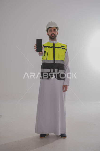 عرض شاشة جوال سوداء فارغة، العمل في القطاع الهندسي، الإعمار بسواعد أبناء الوطن، مفهوم الهندسة والانشاءات المعمارية، بورتريه لمهندس عربي خليجي سعودي يرتدي الثوب التقليدي والسترة الخاصة وخوذة الحماية يقف ويمسك الهاتف المحمول، الجسم بالطول الكامل، خلفية رمادية