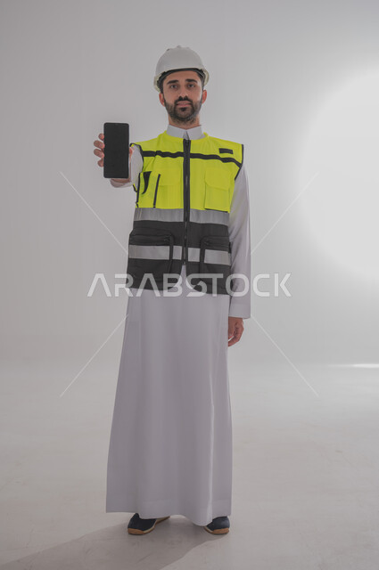 Empty black mobile screen, working in engineering sector, construction by the hands of the sons of the homeland, concept of engineering and architectural construction, portrait of a Saudi Arabian Gulf engineer wearing traditional dress, special jacket and protective helmet standing and holding mobile phone, full body, gray background