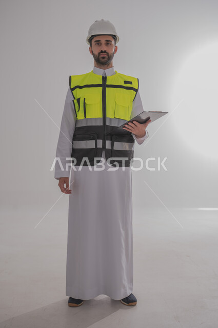 استخدام التقنيات والتكنولوجيا الحديثة في القطاع الهندسي، الإعمار بسواعد أبناء الوطن، مفهوم الهندسة والانشاءات المعمارية، بورتريه لمهندس عربي خليجي سعودي يرتدي الثوب التقليدي والسترة الخاصة وخوذة الحماية يقف ويمسك بجهاز التابلت، الجسم بالطول الكامل، خلفية رمادية