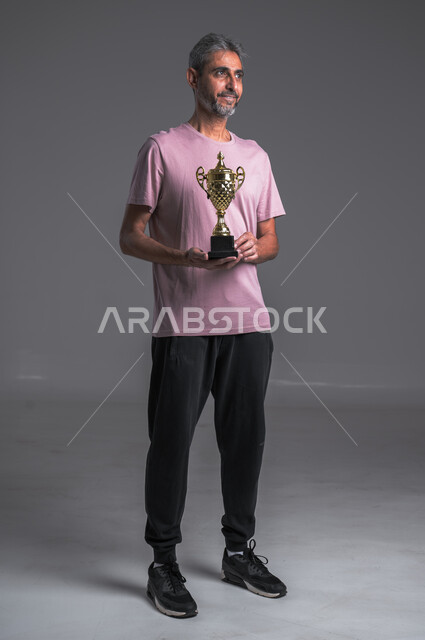 لاعب كرة قدم محترف ،ايماءات وجه ويدين تدل على السعادة والربح ، بورتريه لرجل عربي خليجي سعودي يرتدي الزي الرياضي يحمل بيده كاس الفوز، هواية اللعب بالكرة ، صوره الجسم بالطول الكامل ، خلفيه رمادية