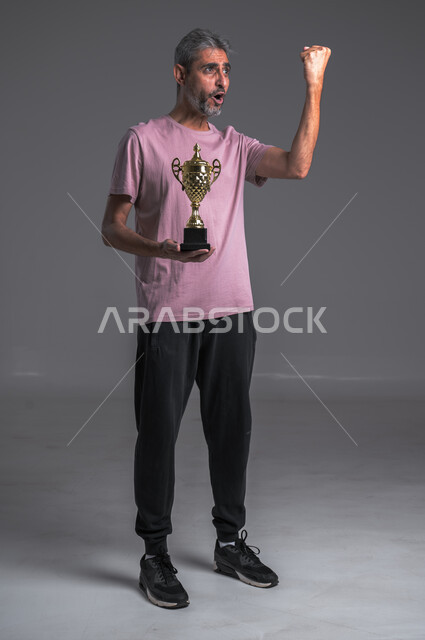ايماءات وجه ويدين تدل على السعادة والربح ،لاعب كرة قدم محترف ، بورتريه لرجل عربي خليجي سعودي يرتدي الزي الرياضي يحمل بيده كاس الفوز، هواية اللعب بالكرة ، صوره الجسم بالطول الكامل ، خلفيه رمادية