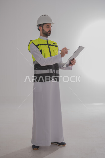 المتابعة والإشراف على العمل بشكل مباشر، الإعمار بسواعد أبناء الوطن، مفهوم الهندسة والانشاءات المعمارية، بورتريه لمهندس عربي خليجي سعودي يرتدي الثوب التقليدي والسترة الخاصة وخوذة الحماية يقف ويقوم بتسجيل الملاحظات على المخطط، الجسم بالطول الكامل، خلفية رمادية