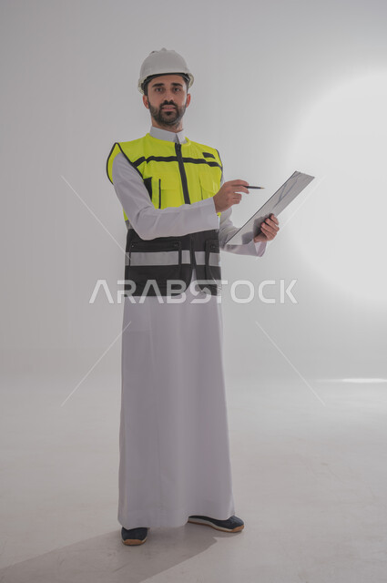 المتابعة والإشراف على العمل بشكل مباشر، الإعمار بسواعد أبناء الوطن، مفهوم الهندسة والانشاءات المعمارية، بورتريه لمهندس عربي خليجي سعودي يرتدي الثوب التقليدي والسترة الخاصة وخوذة الحماية يقف ويقوم بتسجيل الملاحظات على المخطط، الجسم بالطول الكامل، خلفية رمادية