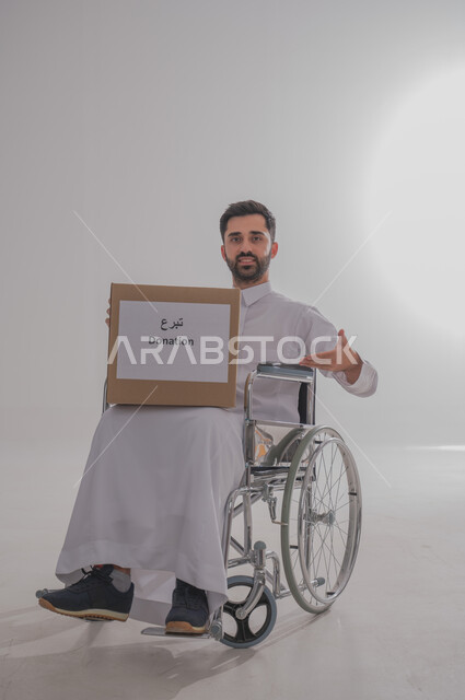 المساعدات والتبرعات الإنسانية، التشجيع على القيام بالأعمال الخيرية الإسلامية، الاحساس بالآخرين ومساعدتهم، بورتريه لشاب عربي خليجي سعودي من ذوي الاحتياجات الخاصة يرتدي الثوب التقليدي يجلس على كرسي متحرك يحمل بيده صندوق تبرع، الجسم بالطول الكامل، خلفية رمادية