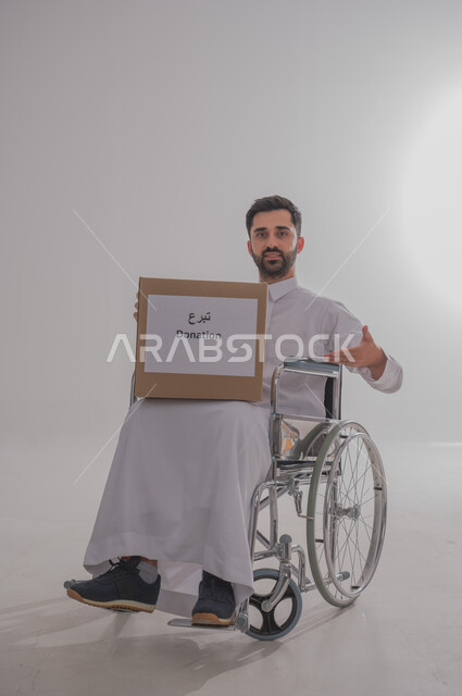 المساعدات والتبرعات الإنسانية، التشجيع على القيام بالأعمال الخيرية الإسلامية، الاحساس بالآخرين ومساعدتهم، بورتريه لشاب عربي خليجي سعودي من ذوي الاحتياجات الخاصة يرتدي الثوب التقليدي يجلس على كرسي متحرك يحمل بيده صندوق تبرع، الجسم بالطول الكامل، خلفية رمادية
