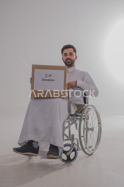 المساعدات والتبرعات الإنسانية، التشجيع على القيام بالأعمال الخيرية الإسلامية، الاحساس بالآخرين ومساعدتهم، بورتريه لشاب عربي خليجي سعودي من ذوي الاحتياجات الخاصة يرتدي الثوب التقليدي يجلس على كرسي متحرك يحمل بيده صندوق تبرع، الجسم بالطول الكامل، خلفية رمادية