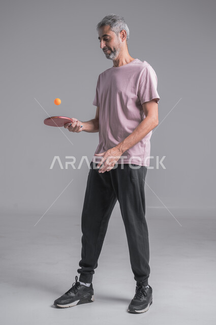 ايماءات الحماس والاندماج، تدريبات وتمرينات رياضيه ،احتراف وإتقان لعبة تنس الطاوله ، رياضات شبابية وأنشطة ترفيهية، بورتريه لرجل عربي خليجي سعودي يضرب الكرة بالمضرب، صوره الجسم بالطول الكامل ، خلفية رماديه