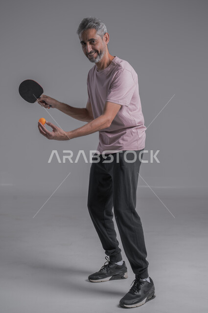 ايماءات  الحماس والاندماج، تدريبات وتمرينات رياضيه ،احتراف وإتقان لعبة تنس الطاوله ، رياضات شبابية وأنشطة ترفيهية، بورتريه لرجل عربي خليجي سعودي يضرب الكرة بالمضرب، صوره الجسم بالطول الكامل ، خلفية رماديه