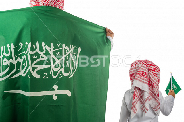 تصوير من الخلف لرجل عربي خليجي سعودي يضع على ظهره علم المملكة العربية السعودية ، طفل خليجي سعودي بيده علم المملكة ،  ذكرى اليوم الوطني السعودي ،الاحتفال بالعيد الوطني السعودي