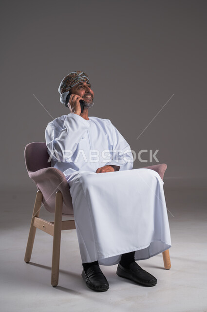 التواصل مع الاهل و الاصدقاء, ايماءات السعادة و السرور، بورتريه  لرجل عربي خليجي عماني  يرتدي الدشداشه و الكمه يجلس على كرسي مريح يقوم بإجراء مكالمة هاتفية عبر الهاتف المحمول، استخدام التقنيات الحديثة المتطورة، صورة الجسم بالكامل, خلفية رمادية