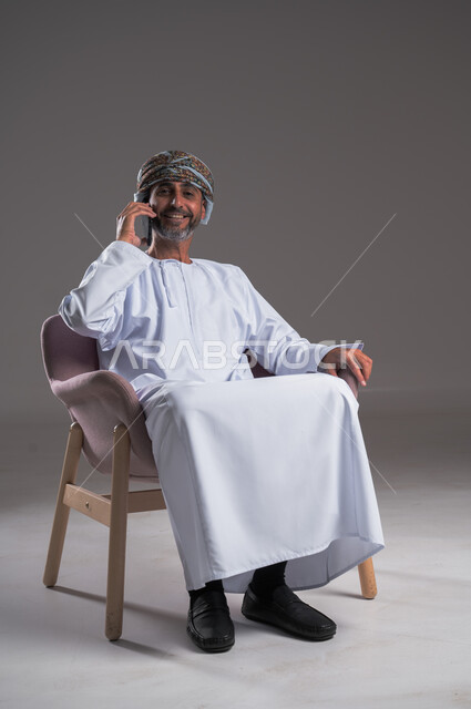 Communication with family and friends, gestures of happiness and pleasure, portrait of an Arab Gulf Omani man wearing a dishdasha and a turban sitting on a comfortable chair making a phone call via a mobile phone, using modern advanced technologies, full body image, gray background
