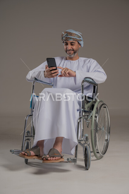 مفهوم صلة الرحم، الاستفادة من التقنيات الحديثة والتكنولوجيا المتطورة، إجراء مكالمات الفيديو باستخدام جهاز الهاتف المحمول،  بورتريه لرجل عربي خليجي عماني مبتسم مقعد يرتدي العمامة والدشداشة يجلس على كرسي متحرك يتواصل مع الأهل والأصدقاء بإيماءات الاندماج والسرور، خلفية رمادية