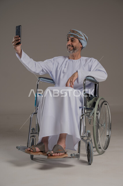 مفهوم صلة الرحم، الاستفادة من التقنيات الحديثة والتكنولوجيا المتطورة، إجراء مكالمات الفيديو باستخدام جهاز الهاتف المحمول،  بورتريه لرجل عربي خليجي عماني مبتسم مقعد يرتدي العمامة والدشداشة يجلس على كرسي متحرك يتواصل مع الأهل والأصدقاء بإيماءات الاندماج والسرور، خلفية رمادية