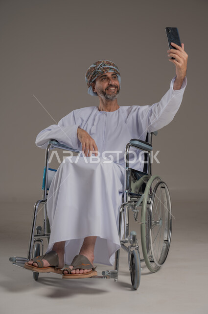 مفهوم صلة الرحم، الاستفادة من التقنيات الحديثة والتكنولوجيا المتطورة، إجراء مكالمات الفيديو باستخدام جهاز الهاتف المحمول،  بورتريه لرجل عربي خليجي عماني مبتسم مقعد يرتدي العمامة والدشداشة يجلس على كرسي متحرك يتواصل مع الأهل والأصدقاء بإيماءات الاندماج والسرور، خلفية رمادية