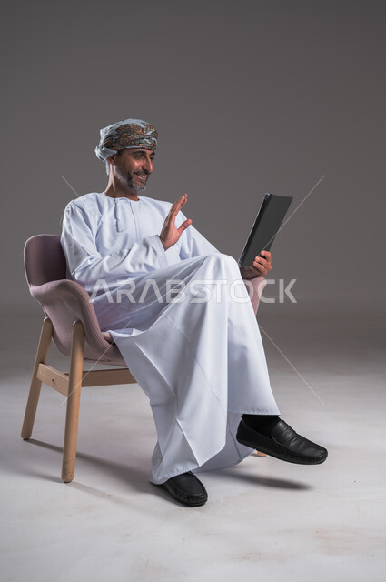 Greeting, welcoming and greeting, taking advantage of modern techniques and advanced technology, making video calls via social networking applications using a tablet, portrait of a smiling Arab Gulf Omani man wearing a turban and dishdasha sitting on a comfortable chair, communicating with family and friends, gray background