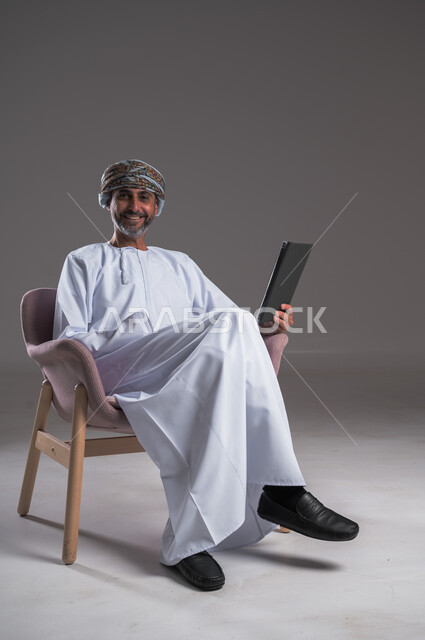 الاستفادة من التقنيات الحديثة والتكنولوجيا المتطورة، إجراء مكالمات الفيديو باستخدام جهاز التابلت،  بورتريه لرجل عربي خليجي عماني مبتسم يرتدي العمامة والدشداشة يجلس على كرسي مريح، التواصل مع الأهل والأصدقاء بإيماءات الاندماج، مفهوم صلة الرحم، خلفية رمادية