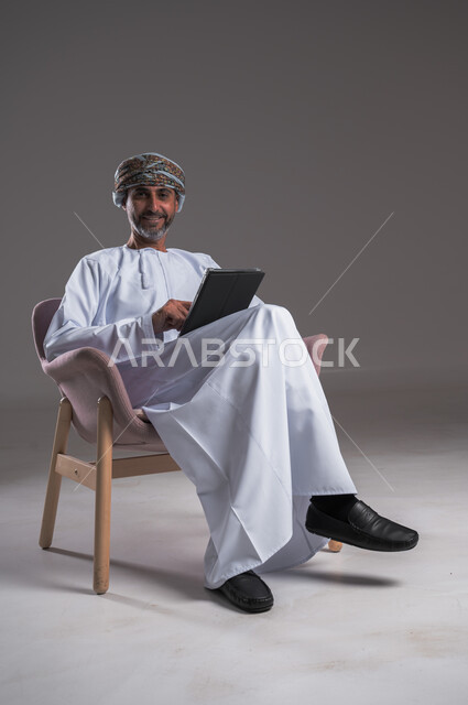 الاستفادة من التقنيات الحديثة والتكنولوجيا المتطورة، إجراء مكالمات الفيديو باستخدام جهاز التابلت،  بورتريه لرجل عربي خليجي عماني مبتسم يرتدي العمامة والدشداشة يجلس على كرسي مريح يتواصل مع الأهل والأصدقاء، النظر الى الكاميرا بإيماءات الفرح، مفهوم صلة الرحم، خلفية رمادية