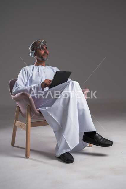 استخدام الوسائل والاجهزة التقنية الحديثة،  بورتريه لرجل عربي خليجي عماني يرتدي العمامة والدشداشة يجلس على كرسي مريح يمسك بيده جهاز التابلت النظر الى مكان ما في الأعلى بإيماءات السرور والسعادة، خلفية رمادية