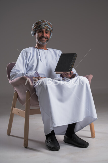 هواية المطالعة والاستكشاف، قضاء وقت الفراغ بالتعلم والثقافة، مفهوم التثقيف والبحث العلمي، بورتريه لرجل عربي خليجي عماني مبتسم يرتدي العمامة والدشداشة يجلس على كرسي مريح يحمل بيده كتاب, النظر الى الكاميرا بإيماءات الفرح والسرور، خلفية رمادية