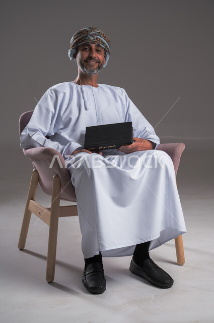 هواية المطالعة والاستكشاف، قضاء وقت الفراغ بالتعلم والثقافة، مفهوم التثقيف والبحث العلمي، بورتريه لرجل عربي خليجي عماني مبتسم يرتدي العمامة والدشداشة يجلس على كرسي مريح يحمل بيده كتاب, النظر الى الكاميرا بإيماءات الفرح والسرور، خلفية رمادية
