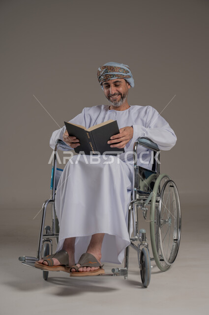 قضاء وقت الفراغ بالتعلم والثقافة، ذوي الاحتياجات الخاصة والهمم، مفهوم التثقيف والبحث العلمي، بورتريه لرجل عربي خليجي عماني مبتسم يرتدي العمامة والدشداشة يجلس على كرسي متحرك يقرأ كتاب بإيماءات الفرح والسرور، خلفية رمادية