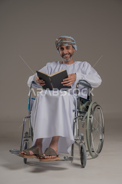 قضاء وقت الفراغ بالتعلم والثقافة، ذوي الاحتياجات الخاصة والهمم، مفهوم التثقيف والبحث العلمي، بورتريه لرجل عربي خليجي عماني مبتسم يرتدي العمامة والدشداشة يجلس على كرسي متحرك يقرأ كتاب, النظر الى الكاميرا بإيماءات الفرح والسرور، خلفية رمادية