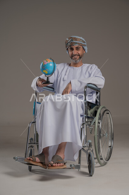 مفهوم البحث والاستكشاف حول العالم، ذوي الاحتياجات الخاصة والهمم، بورتريه لرجل عربي خليجي عماني مبتسم مقعد يرتدي العمامة والدشداشة يجلس على كرسي متحرك يحمل بيده مجسم الكرة الأرضية، خلفية رمادي