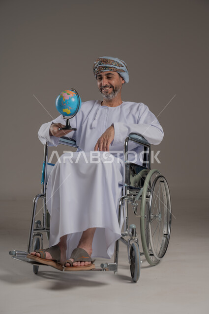 مفهوم البحث والاستكشاف حول العالم، ذوي الاحتياجات الخاصة والهمم، بورتريه لرجل عربي خليجي عماني مبتسم مقعد يرتدي العمامة والدشداشة يجلس على كرسي متحرك يحمل بيده مجسم الكرة الأرضية، خلفية رمادي