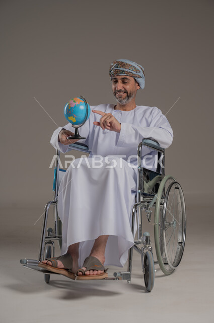 مفهوم البحث والاستكشاف حول العالم، ذوي الاحتياجات الخاصة والهمم، بورتريه لرجل عربي خليجي عماني مبتسم مقعد يرتدي العمامة والدشداشة يحمل بيده مجسم الكرة الأرضية،  الإشارة بإصبع السبابة إلى مكان معين على الخريطة، خلفية رمادية
