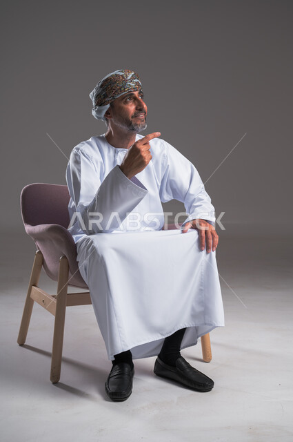 الإشارة والتوضيح إلى شيء ما، بورتريه لرجل عربي خليجي عماني يرتدي العمامة والدشداشة يجلس على كرسي مريح ينظر إلى الاعلى  ويشير بإصبعه السبابة إلى مكان ما، تحديد موقع معين بايماءات السعادة والسرور، إشارات وحركات يدين تدل على هذا المكان (هنا)، خلفية رمادية
