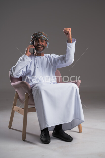 الشعور بالسعادة لسماع خبر ما، تعابير تدل على الفرح والفوز والانتصار، إجراء مكالمات هاتفية عبر الهاتف المحمول, التواصل مع الأهل والأصدقاء، بورتريه لرجل عربي خليجي عماني مبتسم يرتدي العمامة والدشداشة يجلس على كرسي مريح يتكلم على الجوال، خلفية رمادية