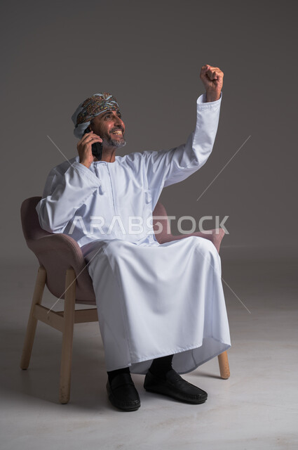 الشعور بالسعادة لسماع خبر ما، تعابير تدل على الفرح والفوز والانتصار، إجراء مكالمات هاتفية عبر الهاتف المحمول, التواصل مع الأهل والأصدقاء، بورتريه لرجل عربي خليجي عماني مبتسم يرتدي العمامة والدشداشة يجلس على كرسي مريح يتكلم على الجوال، خلفية رمادية