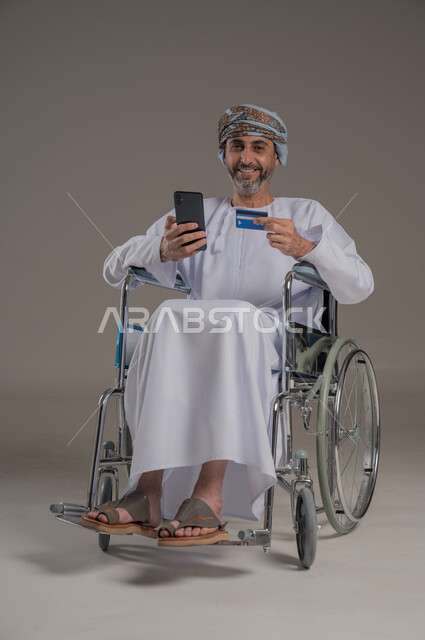الشراء والدفع عن طريق بطاقة الصراف الإئتمانية, بورتريه لرجل عربي خليجي عماني مقعد يرتدي الدشداشة والعمامة يجلس على كرسي متحرك يحمل بيده الهاتف المحمول وكرت البنك المصرفي، سهولة الدفع عبر الحساب البنكي، خلفية رمادية