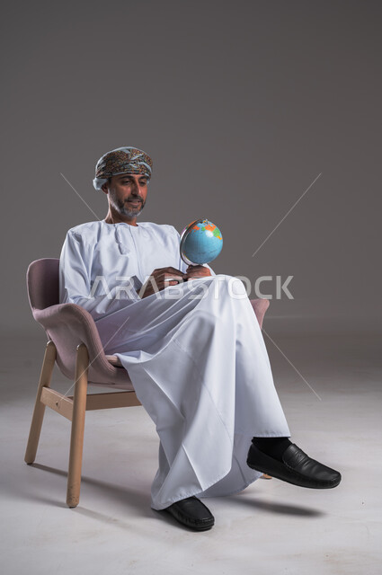 Gestures of concentration and the concept of searching and exploring around the world, looking at one of the continents on the map, portrait of an Arab Gulf Omani man wearing a dishdasha and turban sitting on a comfortable chair holding a globe in his hand, full body image, gray background